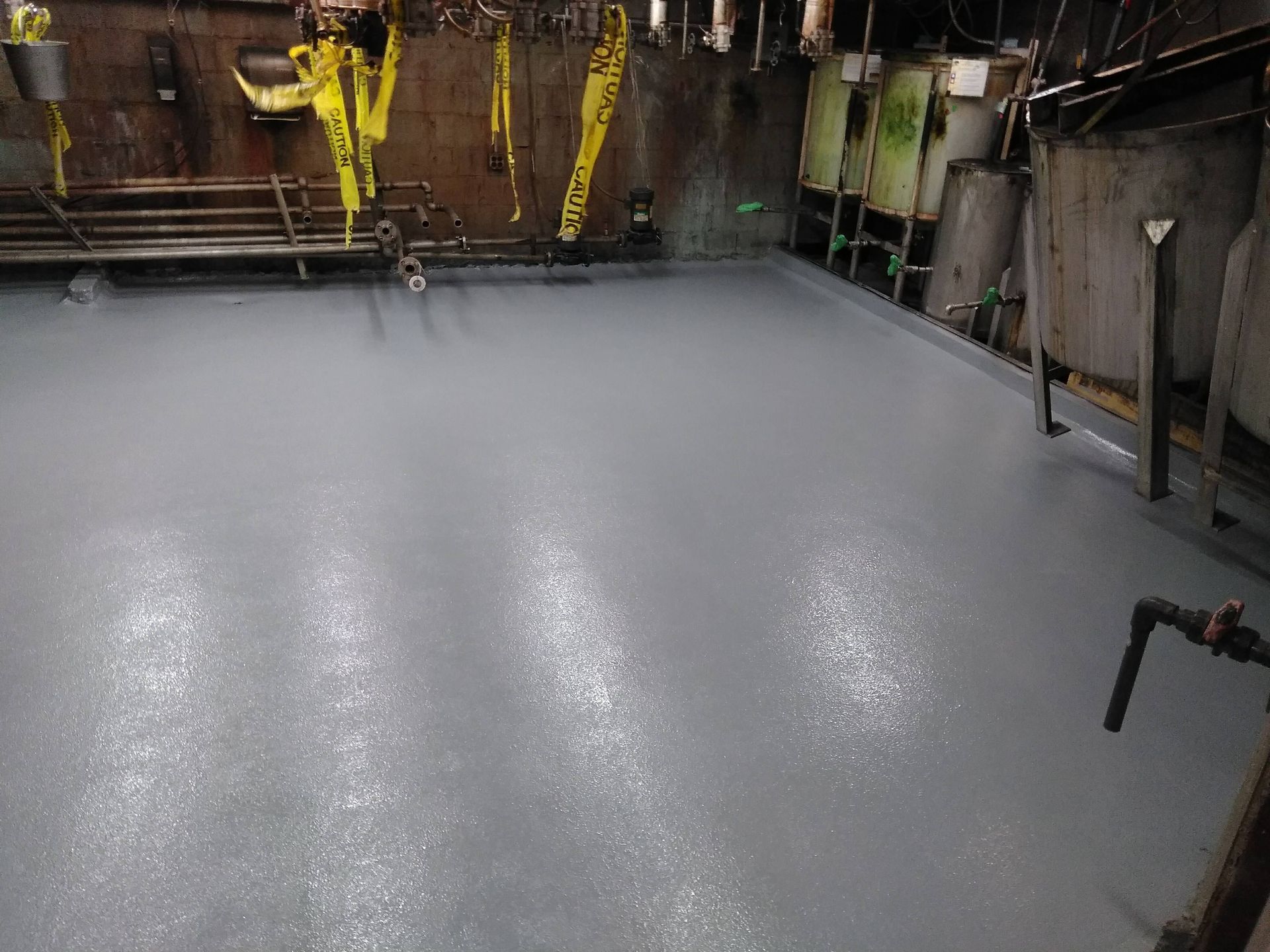 Gray, wet floor in an industrial setting with pipes, walls, and overhead equipment.