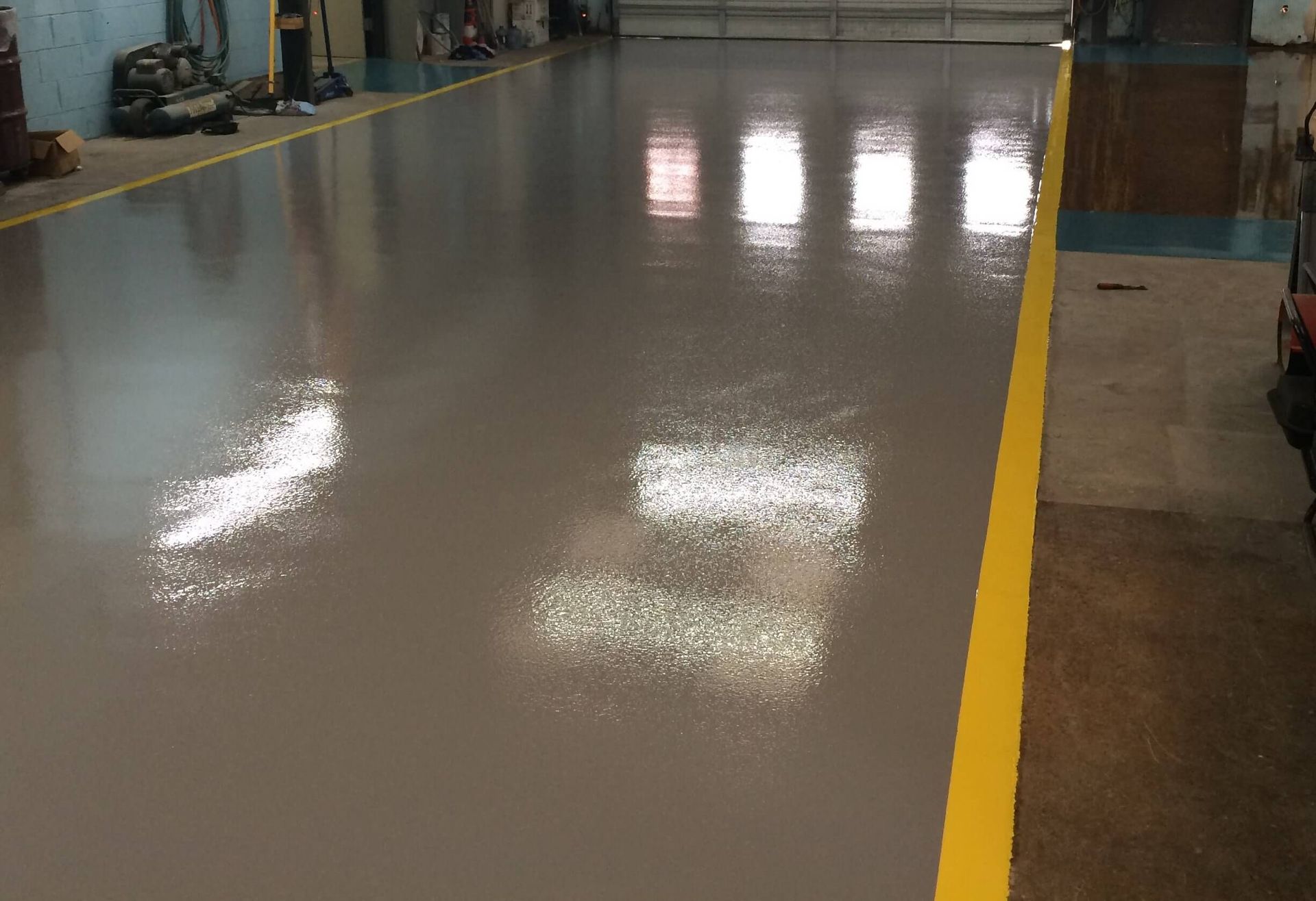 Gray, glossy epoxy floor in a workshop, reflecting bright window light. Yellow line marks the edge.