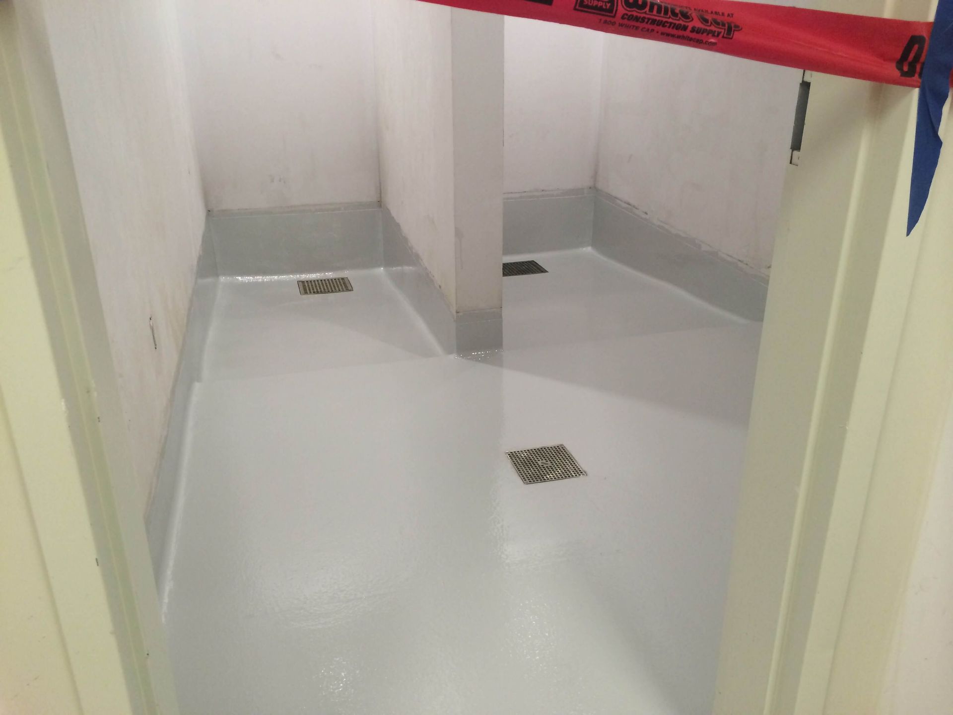 Two stalls with light gray flooring and walls, drain covers. One stall has a red tape across it.