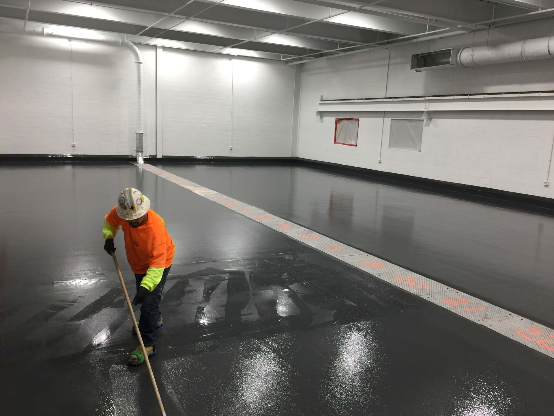 Person in orange shirt and hard hat squeegeeing a shiny, dark gray floor in a large, empty room.