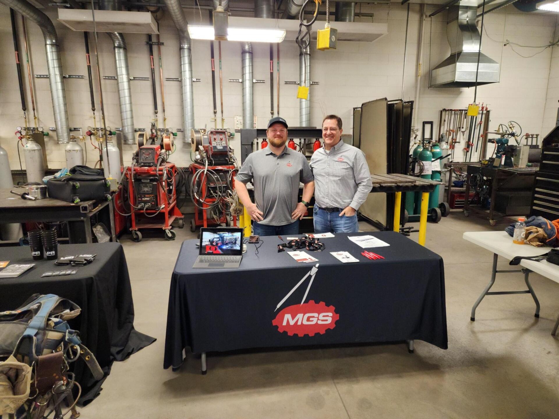 Two men are standing in front of a table with a mgs logo on it.