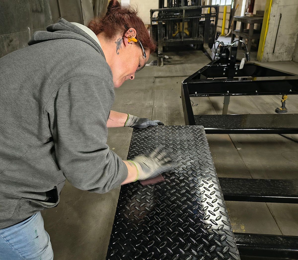 A person in safety glasses and gloves sanding a diamond plate on a black metal frame. Industrial setting.