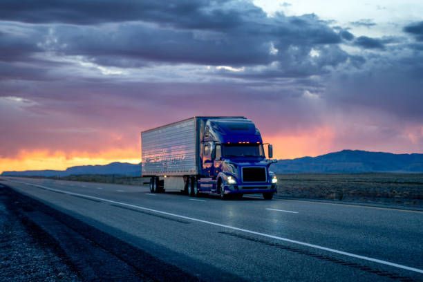 A blue semi truck is driving down a highway at sunset.