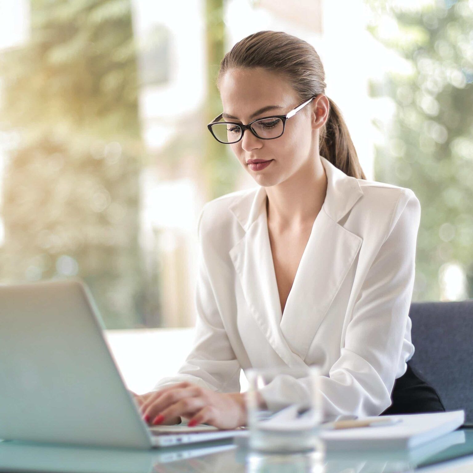 A woman wearing glasses is sitting at a desk using a laptop computer.