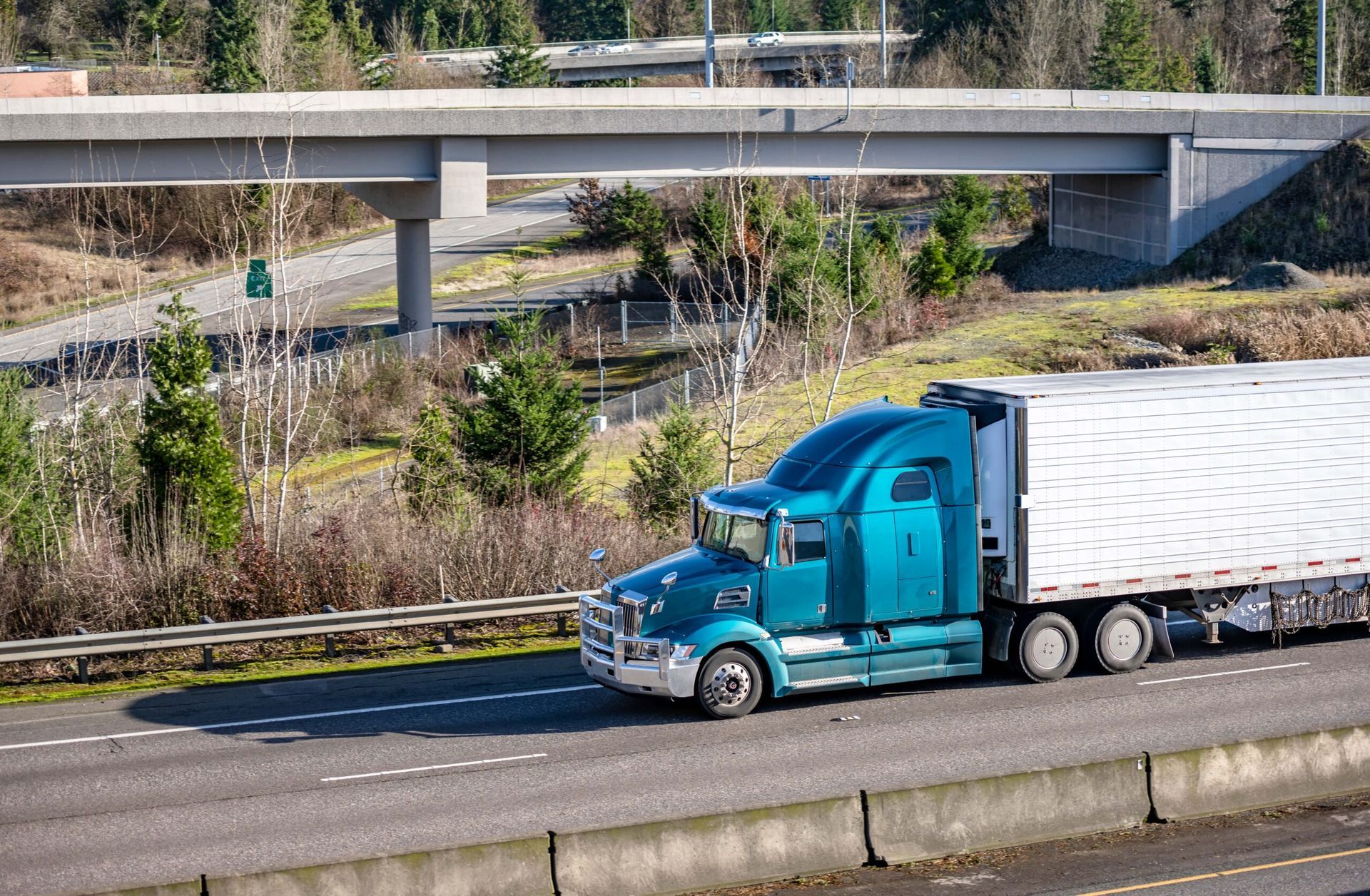 A blue semi truck is driving down a highway under a bridge.