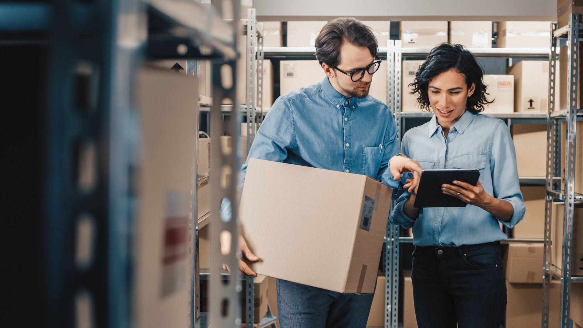 A man is holding a cardboard box and a woman is looking at a tablet in a warehouse.