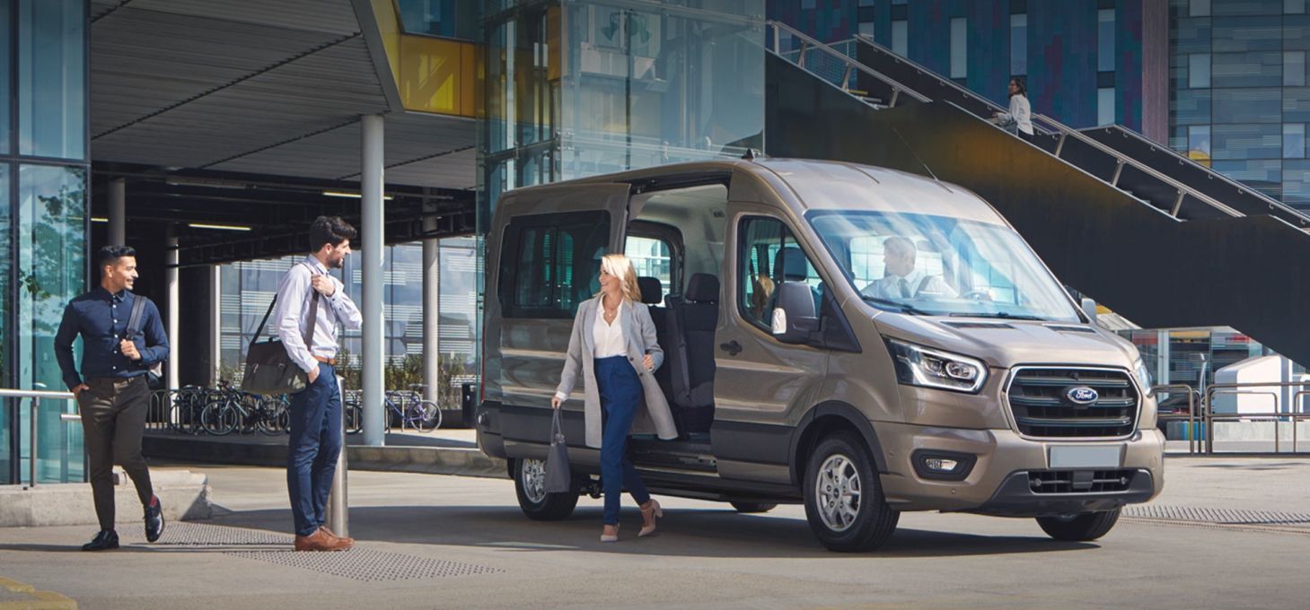People exiting a brown Ford Transit van in front of a modern building.