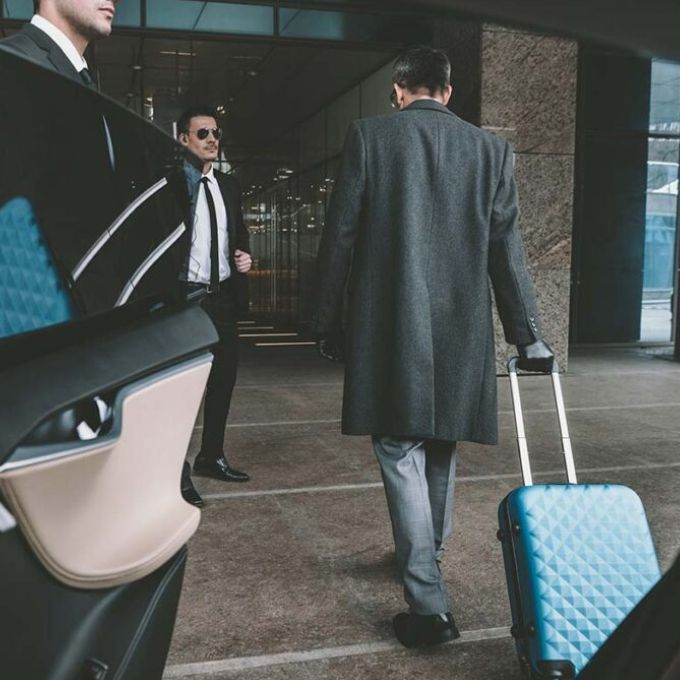 Man in gray coat pulling suitcase, two men in suits near car. Hotel entrance.