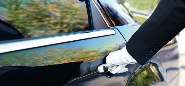 A gloved hand in a dark suit opens the door of a black car.