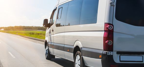 A white passenger van driving on a road with green vegetation on the side and a sunny sky.