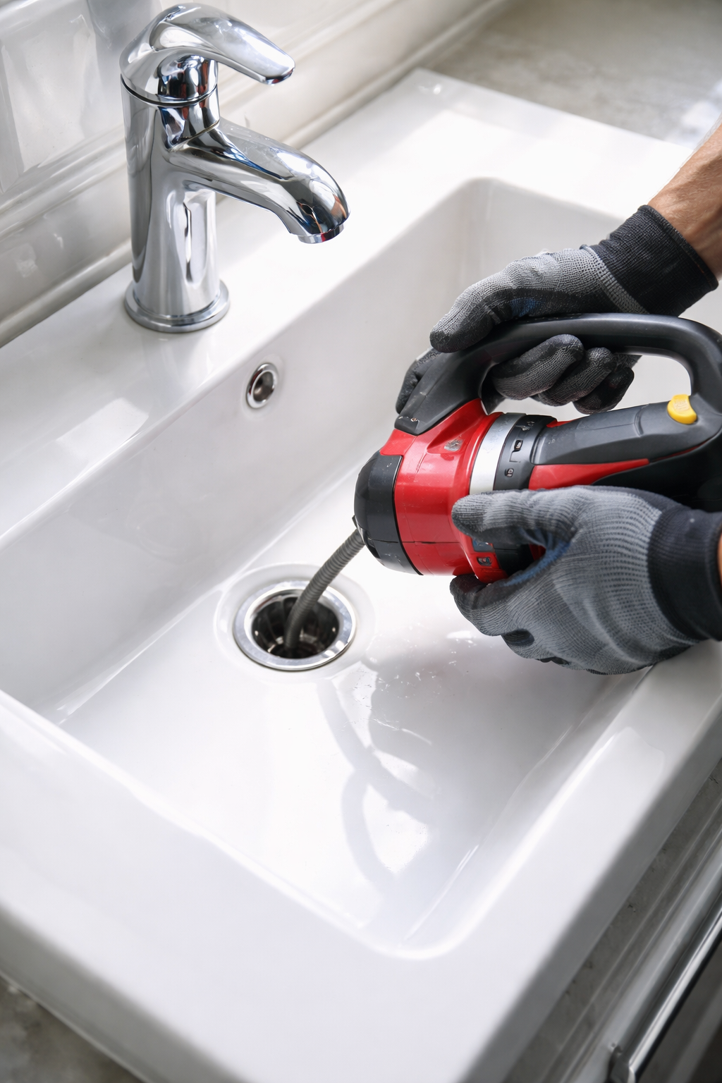 A person wearing grey work gloves uses a red handheld power tool to clear a clog in a bathroom sink drain.