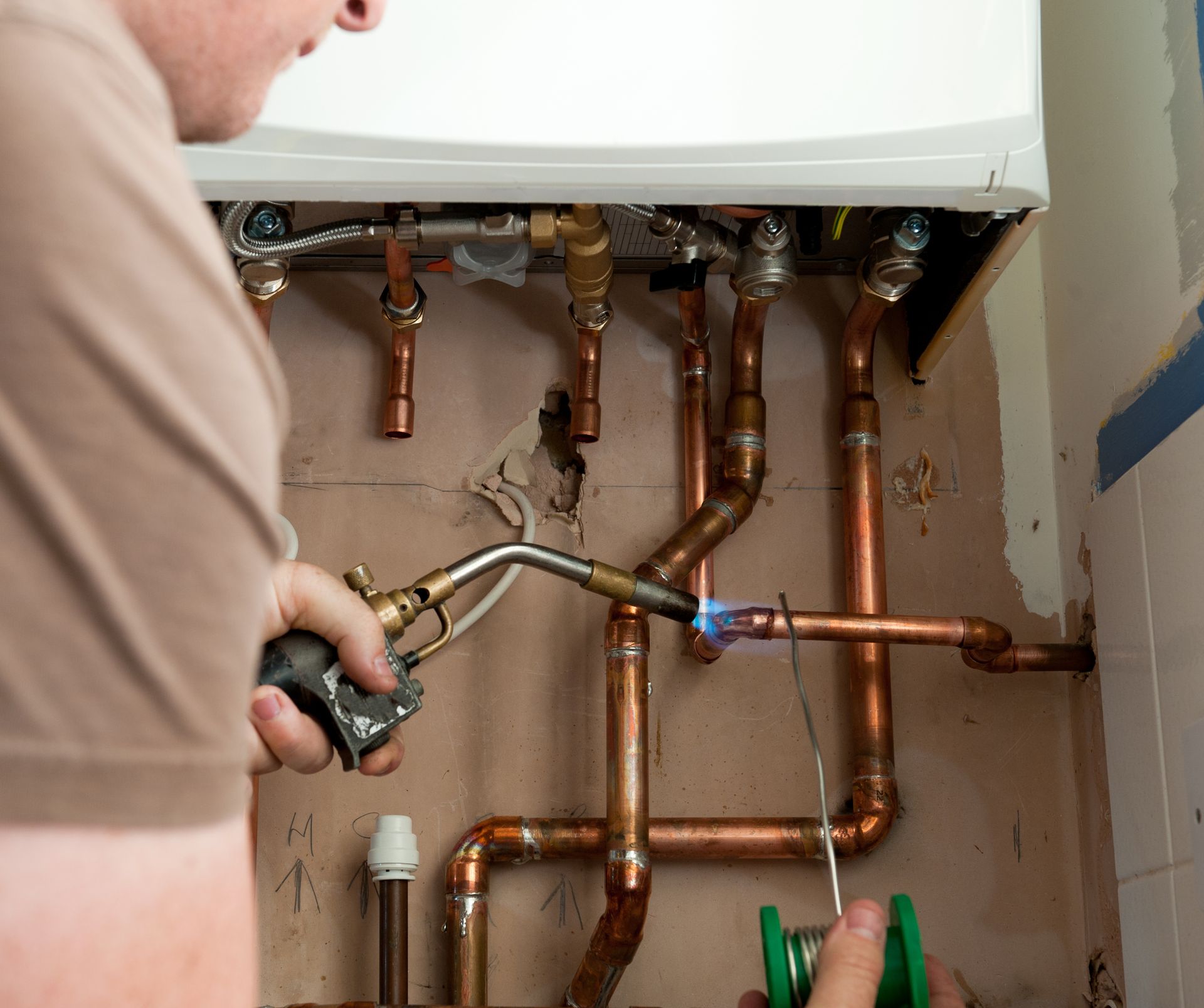A plumber uses a blowtorch to solder copper pipes connected to a wall-mounted boiler.