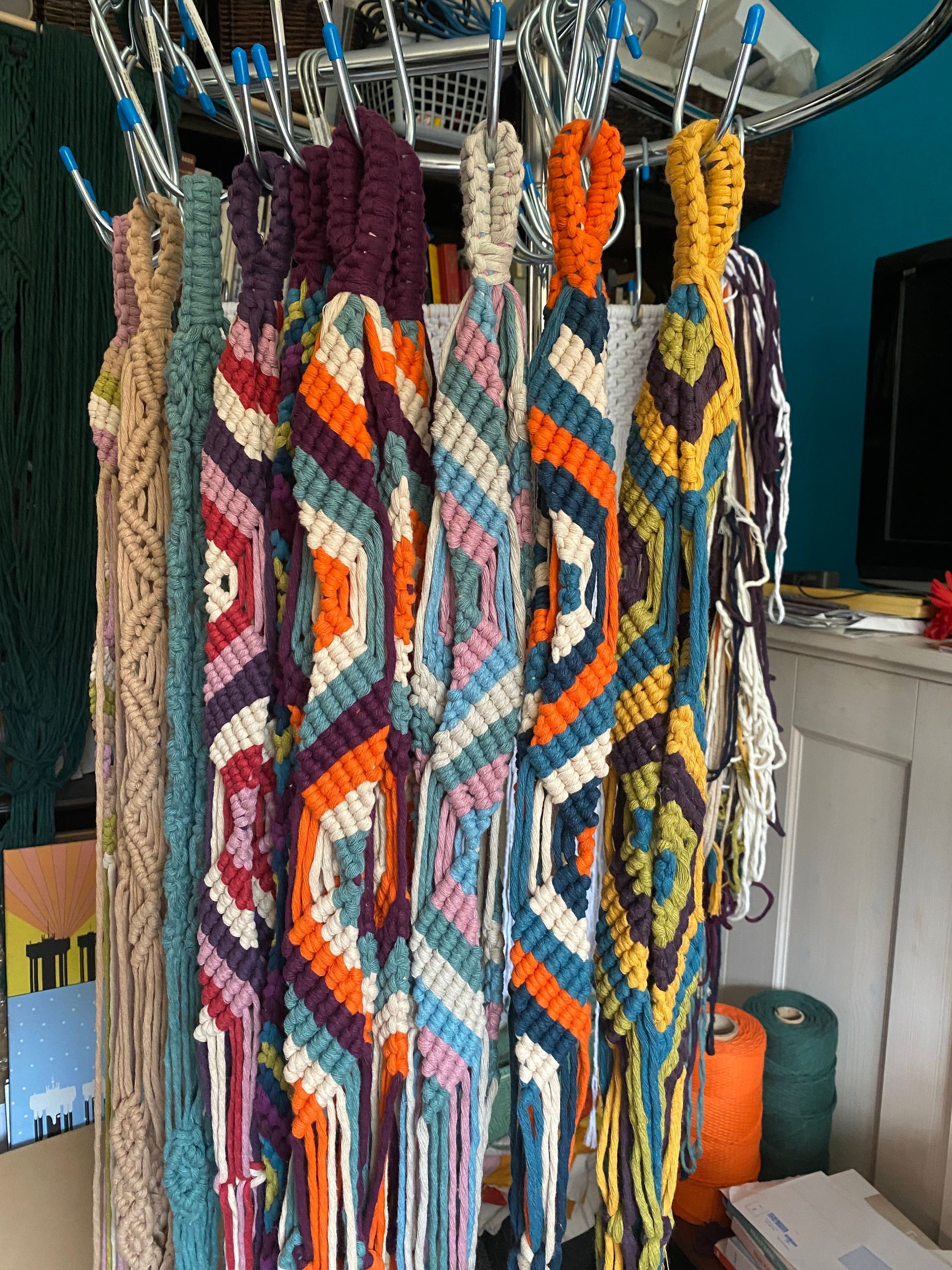A selection of plant hangers, hanging from a rail,  in a variety of colours made using densely knotted half hitches.