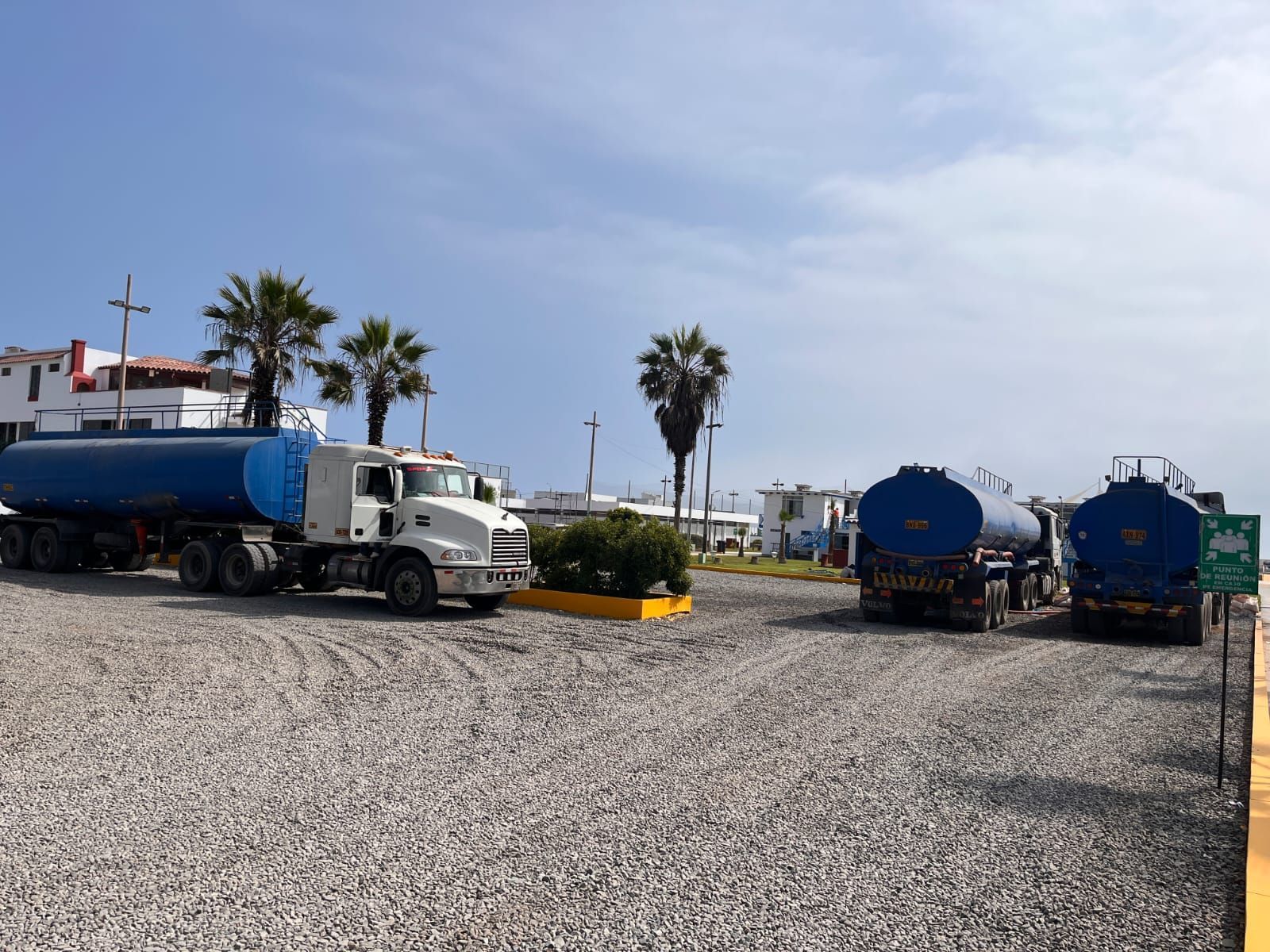 Tres camiones cisterna de agua azul estacionados en un lote de grava cerca de edificios y palmeras bajo un cielo azul.