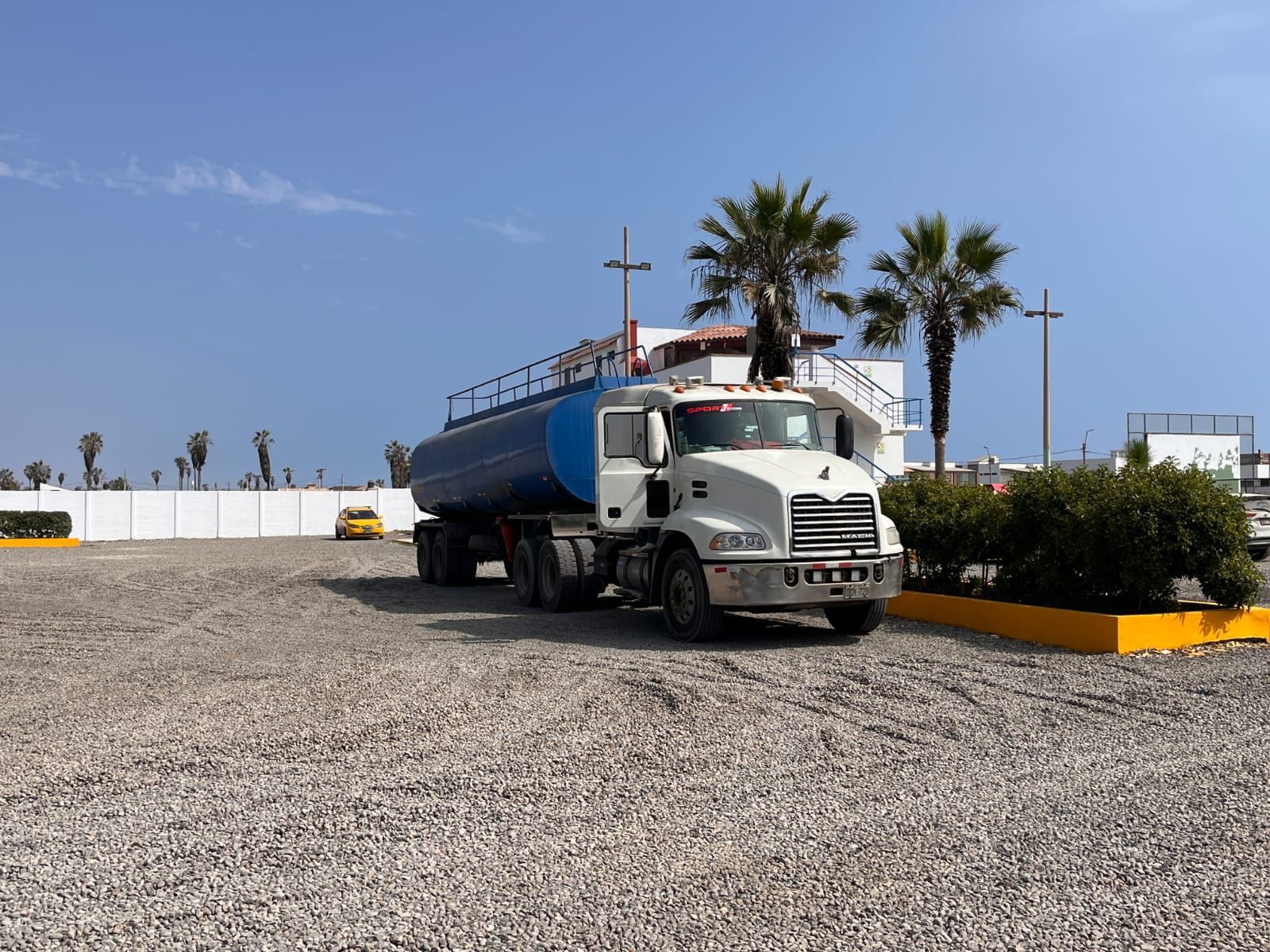 Camión cisterna de agua estacionado en un lote de grava cerca de un edificio blanco y palmeras.