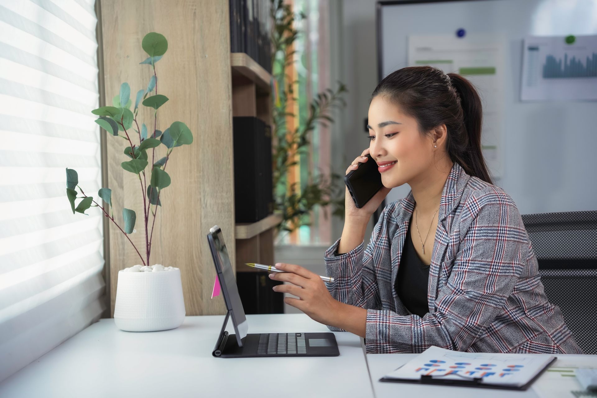 A woman is sitting at a desk talking on a cell phone while looking at a laptop.
