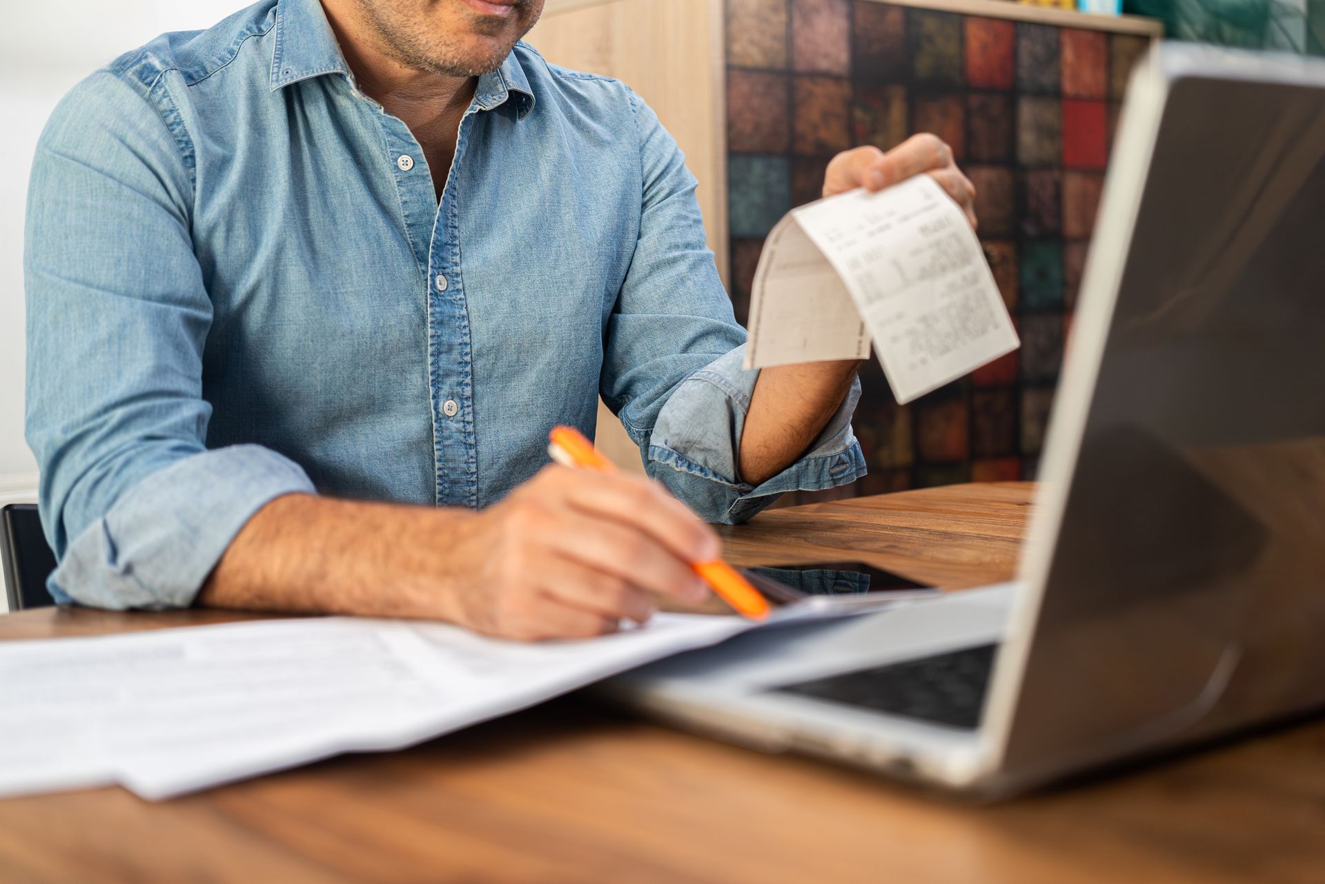A man is sitting at a table holding a receipt in front of a laptop computer.