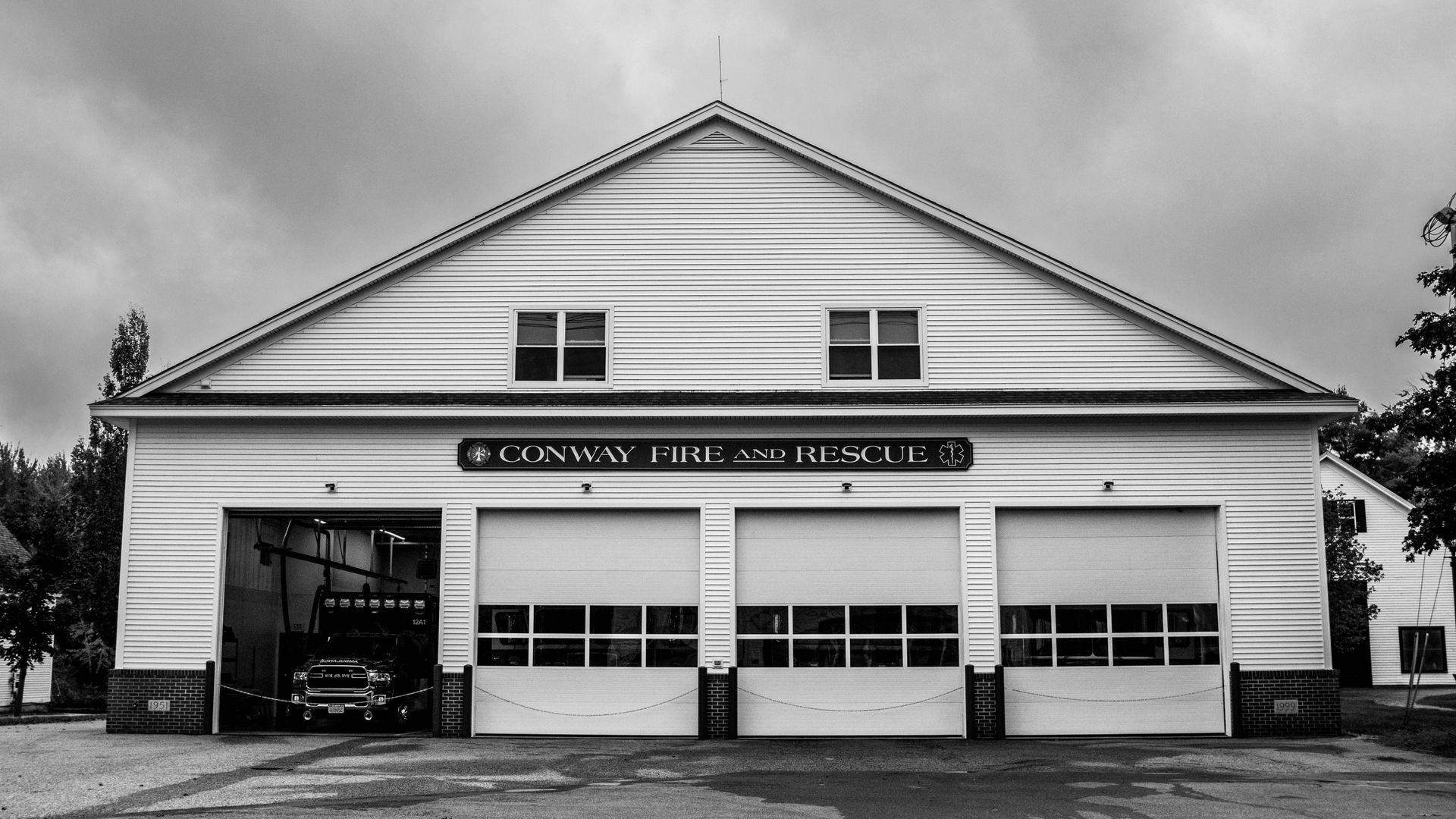 Fire station with three garage doors and a vehicle inside the open left door, under a cloudy sky.
