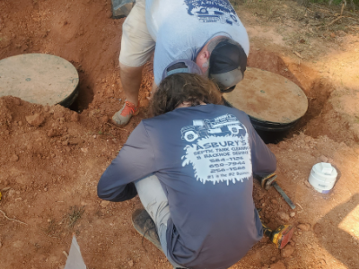 Two people work on a septic tank, kneeling in a dirt pit to inspect two round concrete access covers.