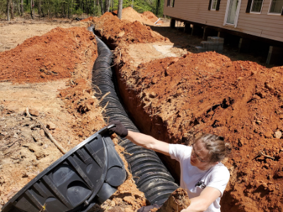 A person installs a black plastic drain pipe in a deep trench beside a house.