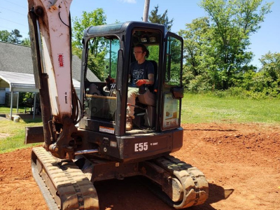 A person sits in the open cab of a Bobcat E55 excavator parked on a dirt lot on a sunny day.