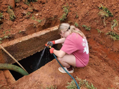 A person in a red shirt and work gloves uses a hose to spray water into an open, excavated concrete utility box.