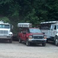 Three utility trucks, including a red pickup and two white flatbed trucks, are parked on a dirt lot near trees.