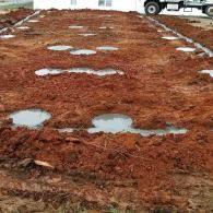 Red dirt construction site with multiple poured concrete foundation footings containing puddles of rainwater.