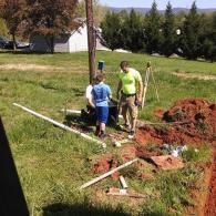 Two people working near a utility pole and an open trench in a grassy, outdoor area on a sunny day.