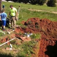 A person in a bright yellow shirt and shorts works near an open trench in a grassy area with a surveying tripod nearby.