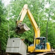 A yellow excavator uses its bucket to lift a large, grey concrete utility box in a wooded area.