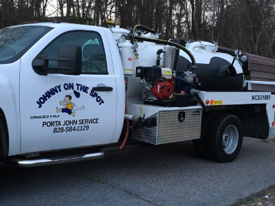 A white Johnny On The Spot porta-john service truck with a black tank, parked outdoors with woods in the background.