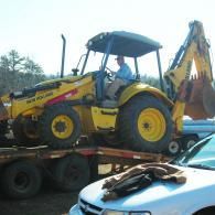 A yellow tractor is loaded onto a flatbed trailer outdoors, with a white car parked in the foreground.