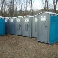 A row of portable grey and blue toilets lined up on a gravel lot in front of a wooded area.