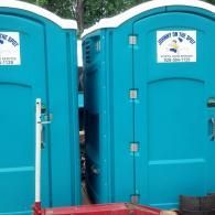 Two bright teal portable toilets sit side-by-side with white logos on the doors.