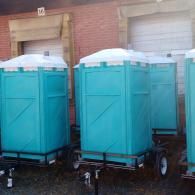 Several teal portable toilets on trailers lined up in front of a brick building with garage doors.