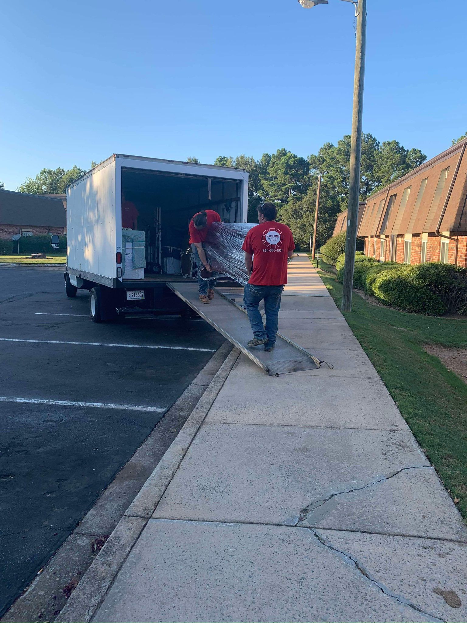 Two people unloading a white truck on a ramp, near a sidewalk and buildings.