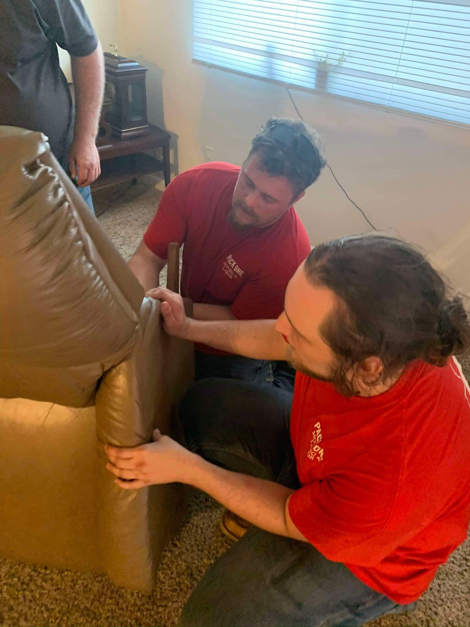 Two men in red shirts assembling a tan leather couch, indoors. A third person stands nearby.