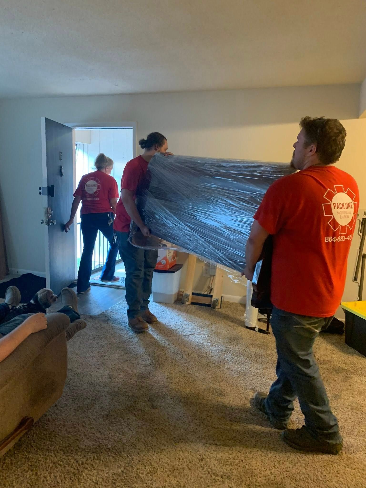 Movers carrying a wrapped sofa through a doorway. Three people in red shirts inside a room.