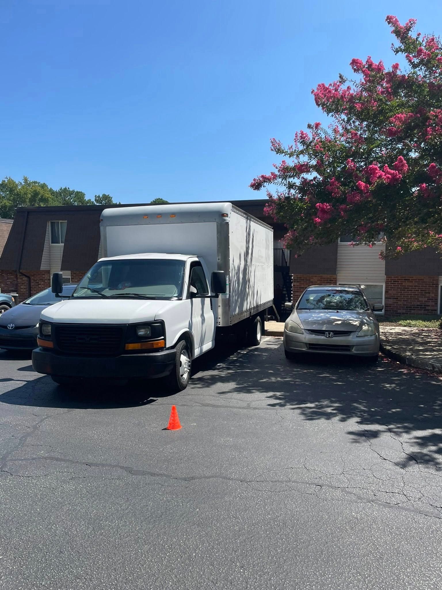 White moving truck parked on asphalt with cars and apartment building in the background. Bright sunny day.