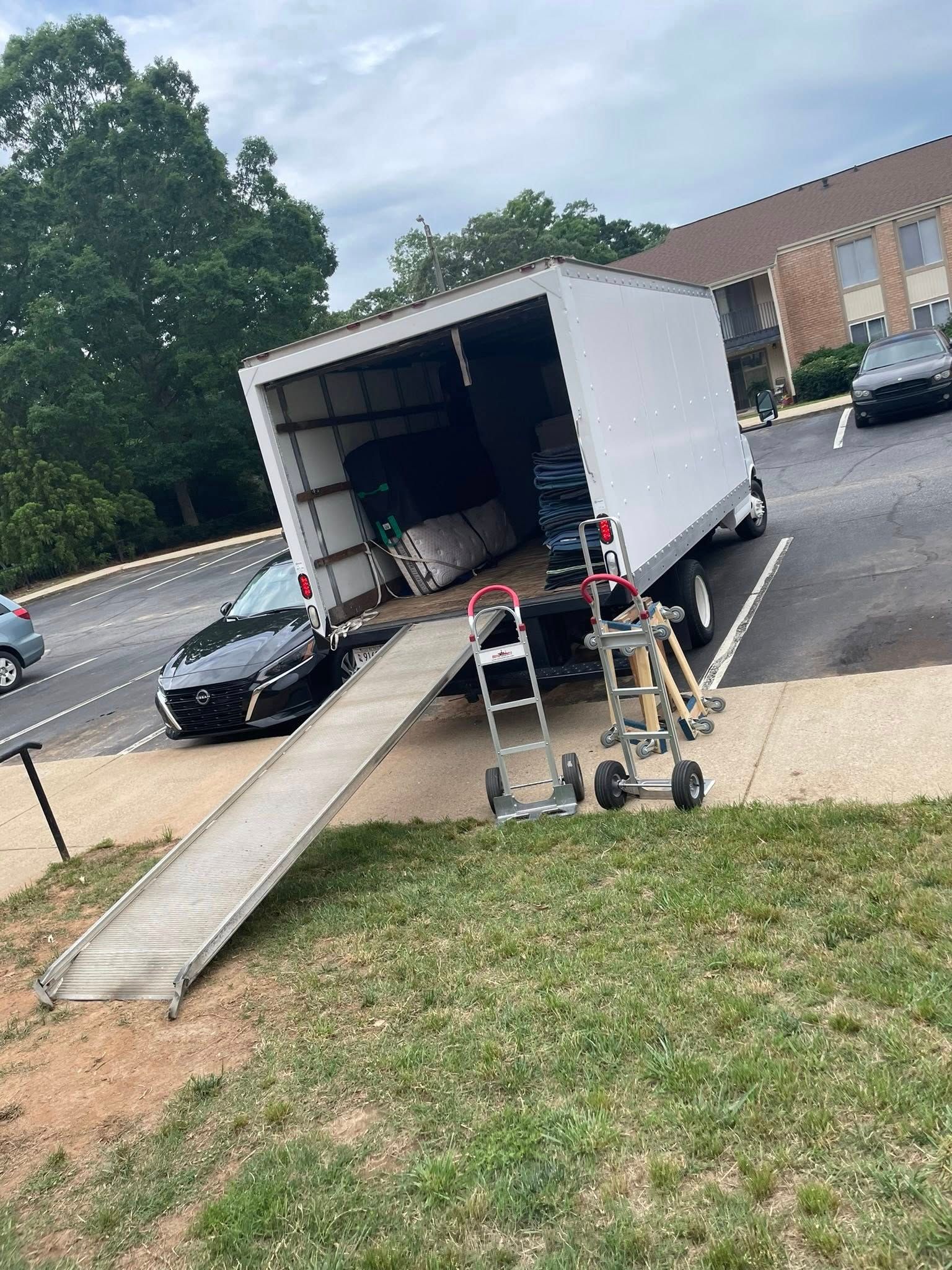 A white moving truck parked with its ramp extended. A black car is parked nearby.