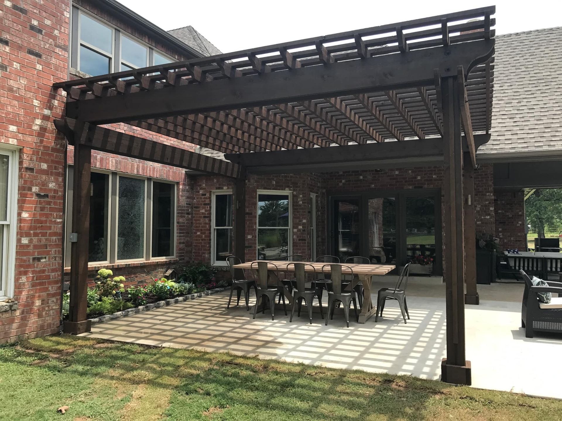 A patio with a table and chairs under a pergola in front of a brick house.