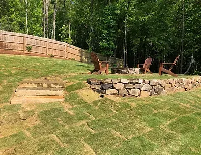 A backyard with a stone wall , chairs and a fire pit.