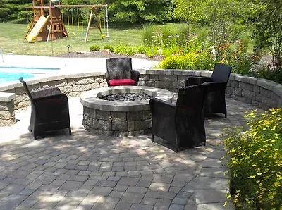 A fire pit is surrounded by chairs on a patio next to a pool.