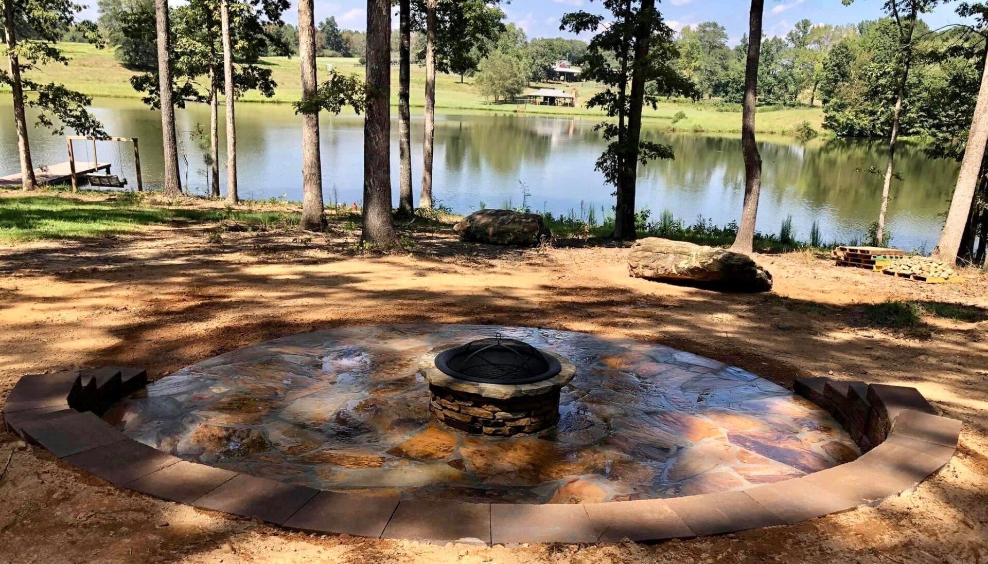 A fire pit is surrounded by trees next to a lake.
