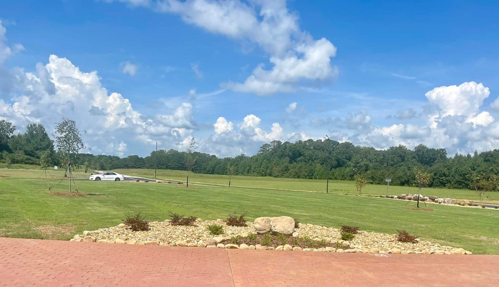 A large grassy field with trees in the background and a red driveway in the foreground.