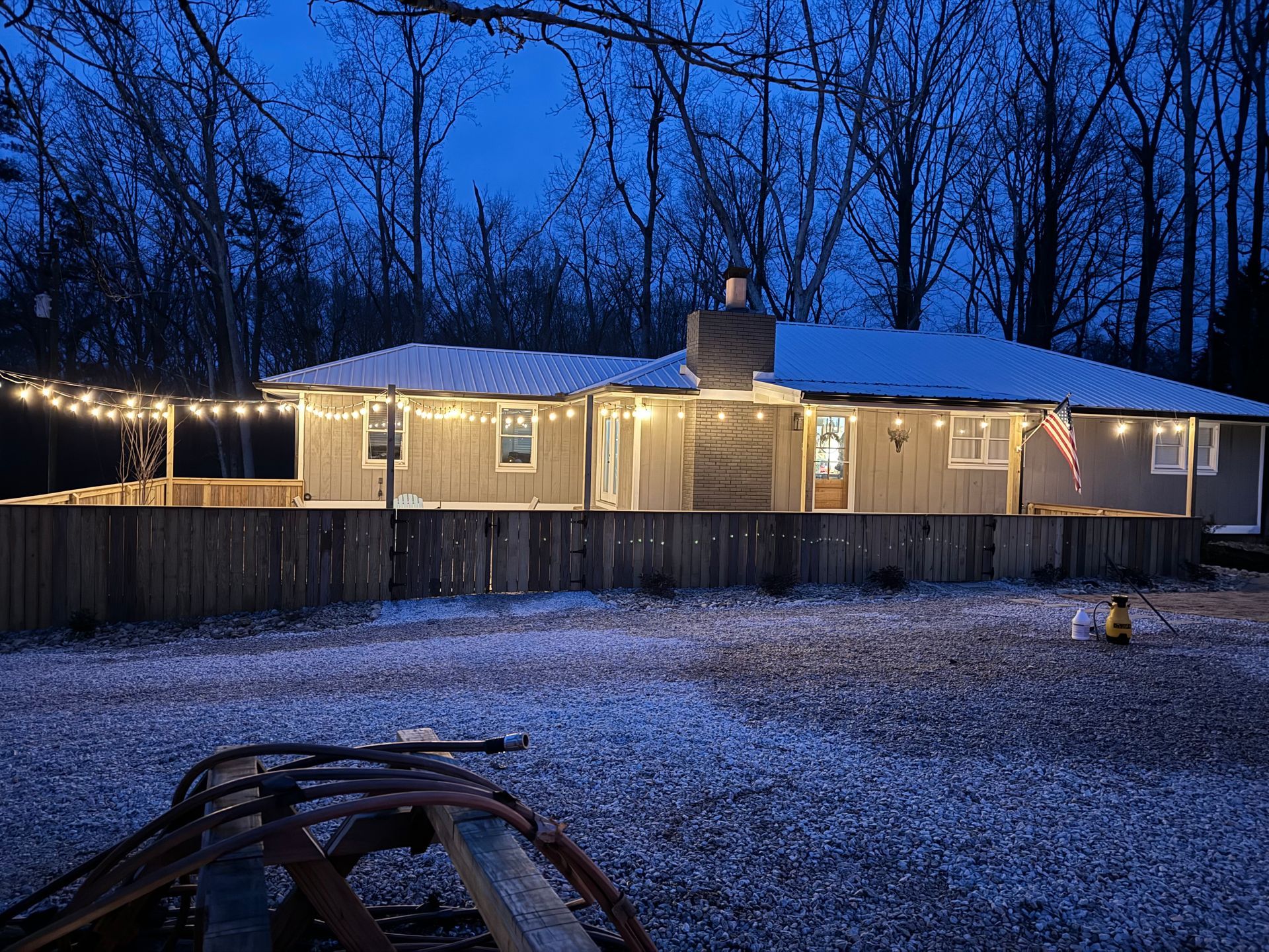 A house at dusk with string lights, a wooden fence, and a snow-covered lawn.
