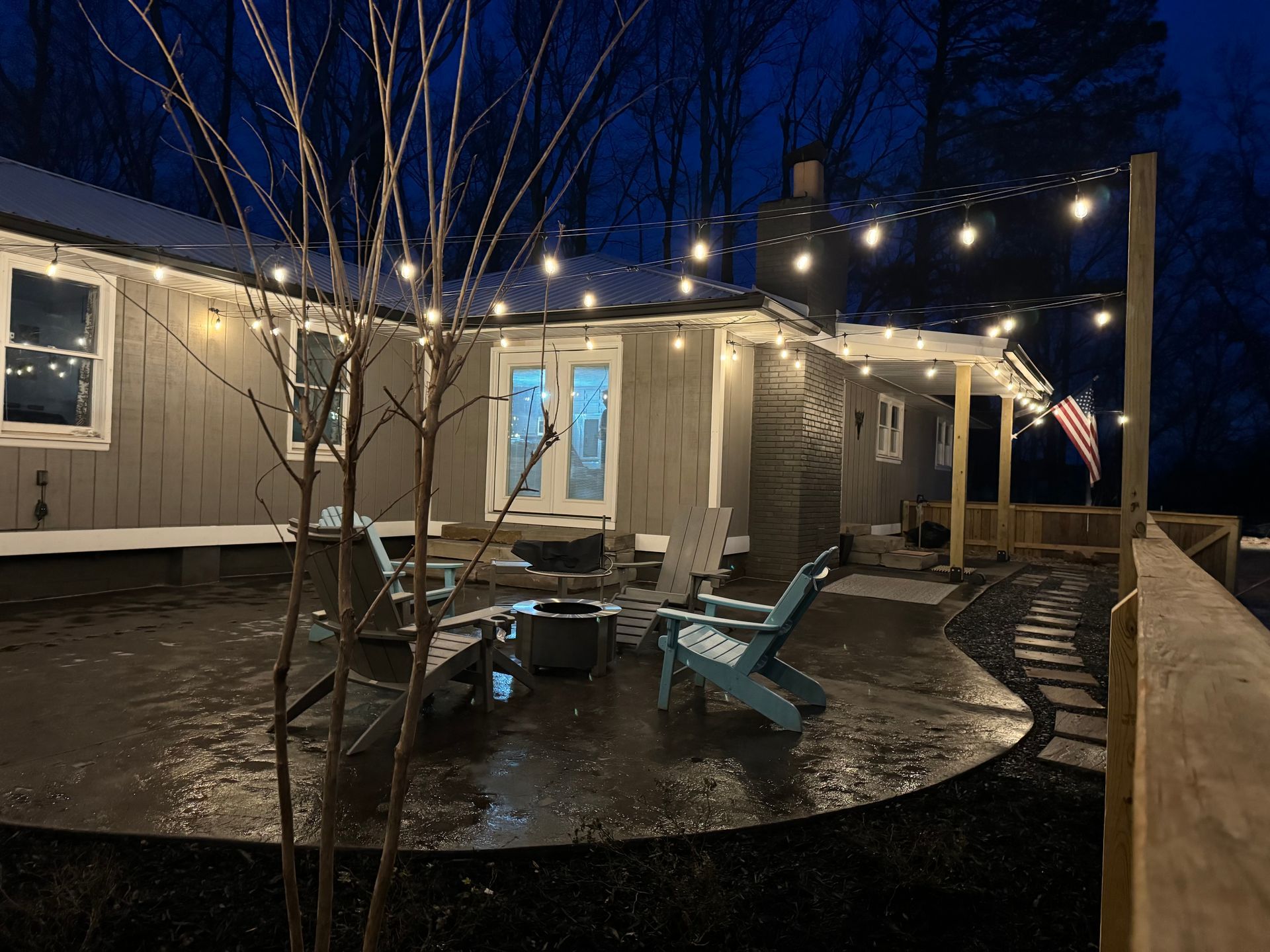 Nighttime backyard with lit string lights, Adirondack chairs, and a fire pit near a house.
