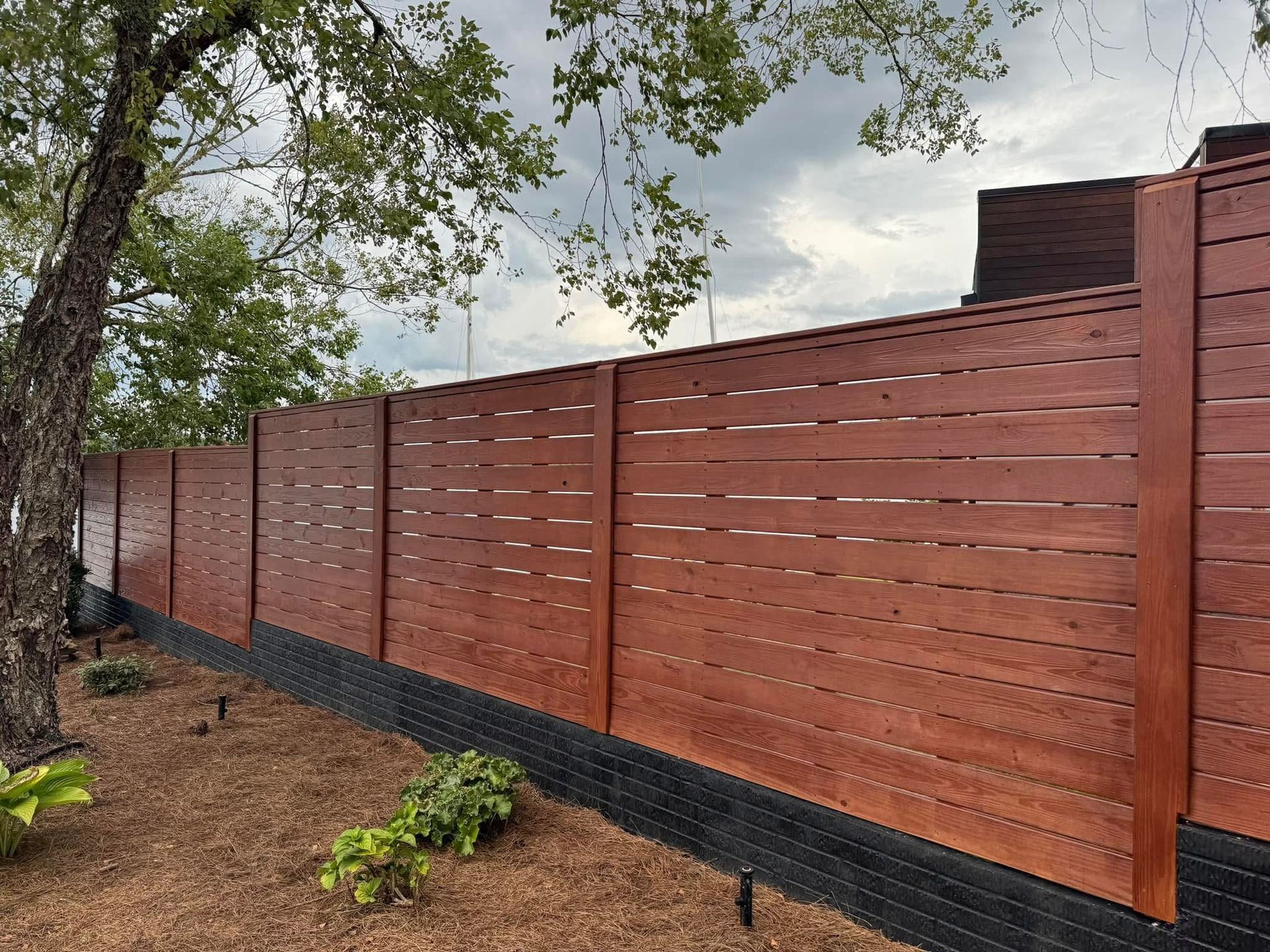 Brown wooden horizontal slat fence along a yard with a tree and overcast sky.