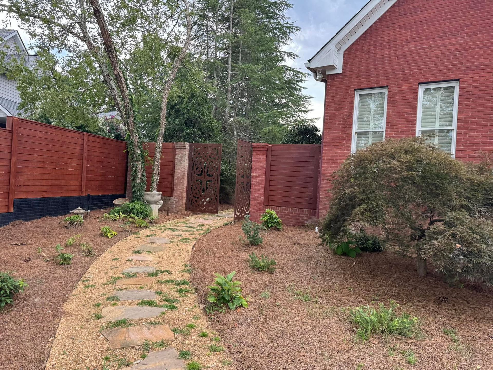 Stone pathway leads to a decorative metal gate and wooden fence next to a red brick house with a garden.